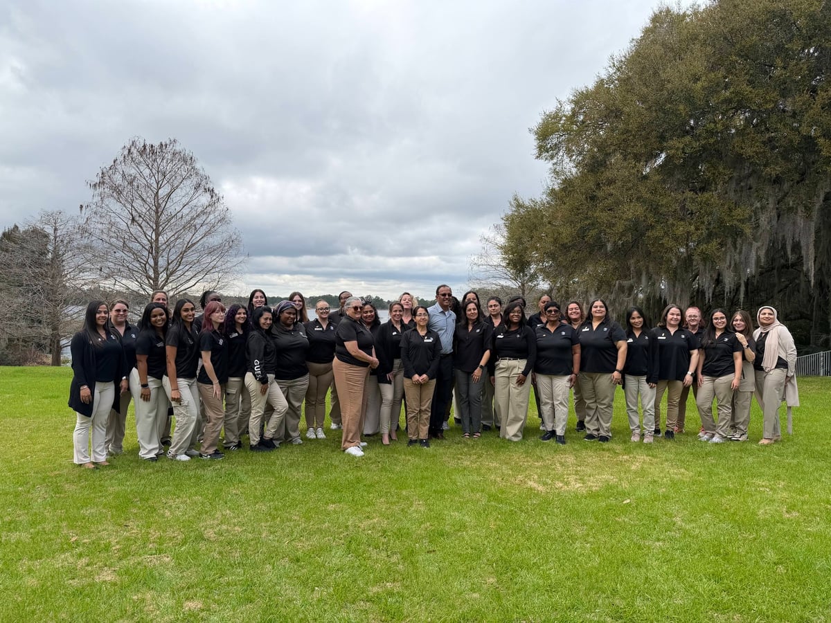 women in khaki pants and back polos standing on grass in front of a lake