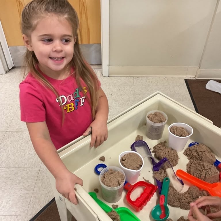 Preschool student using kinetic sand and scooping tools to strengthen fine motor skills, creativity, sensory exploration, and imaginative play at Primrose School.