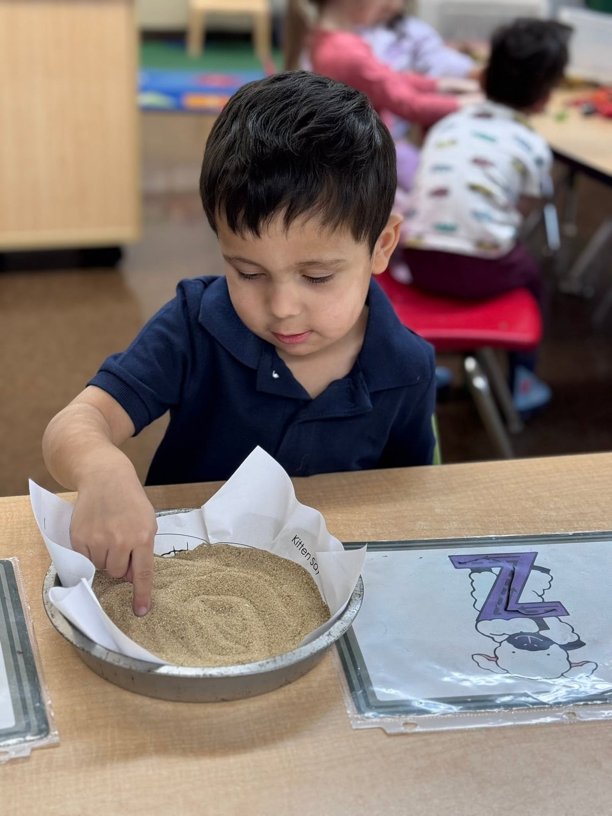 Preschooler tracing letters in sand to strengthen letter recognition, fine motor skills, and early handwriting development during a hands-on literacy activity at Primrose School.
