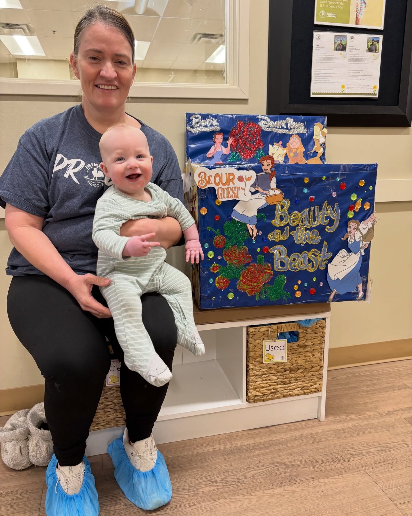 Teacher holding a smiling infant next to a Beauty and the Beast-themed family project display showcasing creativity, family involvement, and classroom learning.