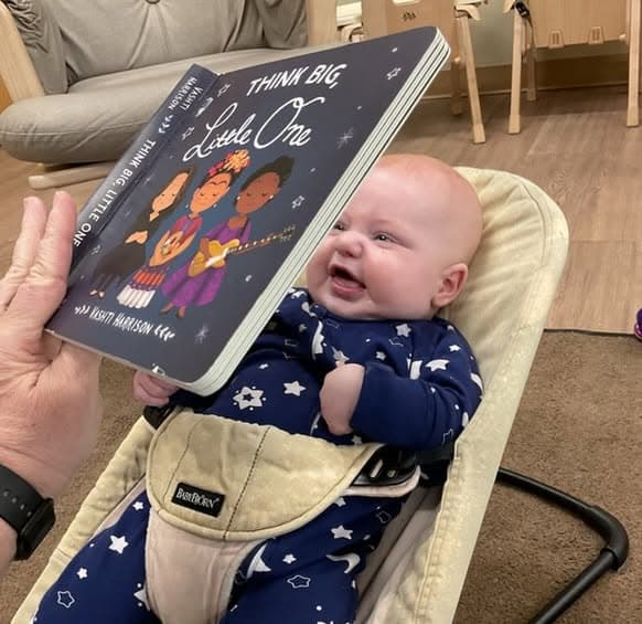 Infant smiling during story time while listening to a teacher read Think Big, Little One, encouraging early literacy and language development.