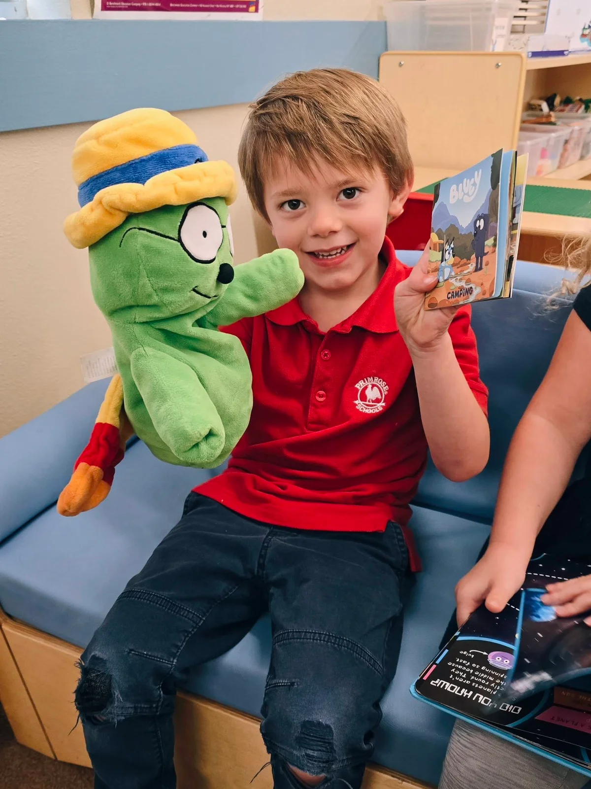 Smiling Pre-K boy holding up a book and showing it to OG the Bookworm at Primrose School at Heritage.