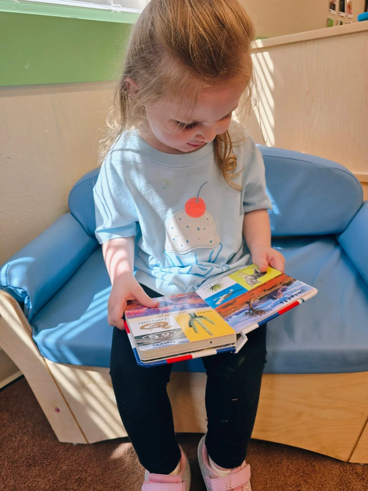 2 year old grl sitting on the couch reading a children's book in a daycare classroom at Primrose School at Heritage.