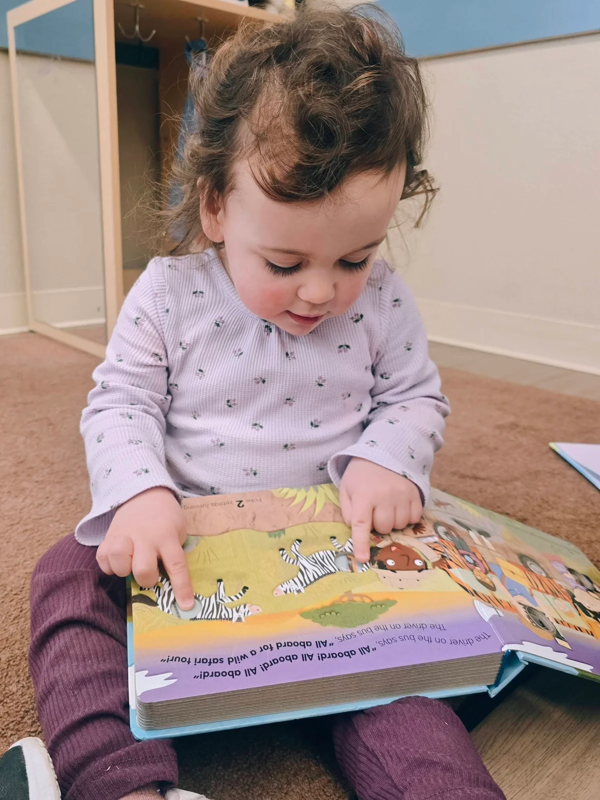 Toddler girl sitting on the floor reading a children's book in a daycare classroom at Primrose School at Heritage.