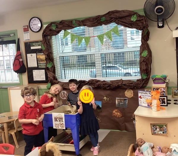 Young children at Primrose School of NE Flower Mound dressed as paleontologists use small paintbrushes to carefully excavate toy dinosaur fossils in a sensory bin alongside a classroom mascot cat, engaging in hands-on STEM learning and dramatic play.