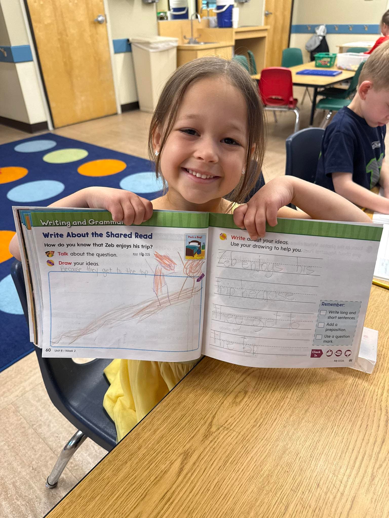 Smiling student proudly holding up a completed writing and grammar workbook page during literacy learning time.
