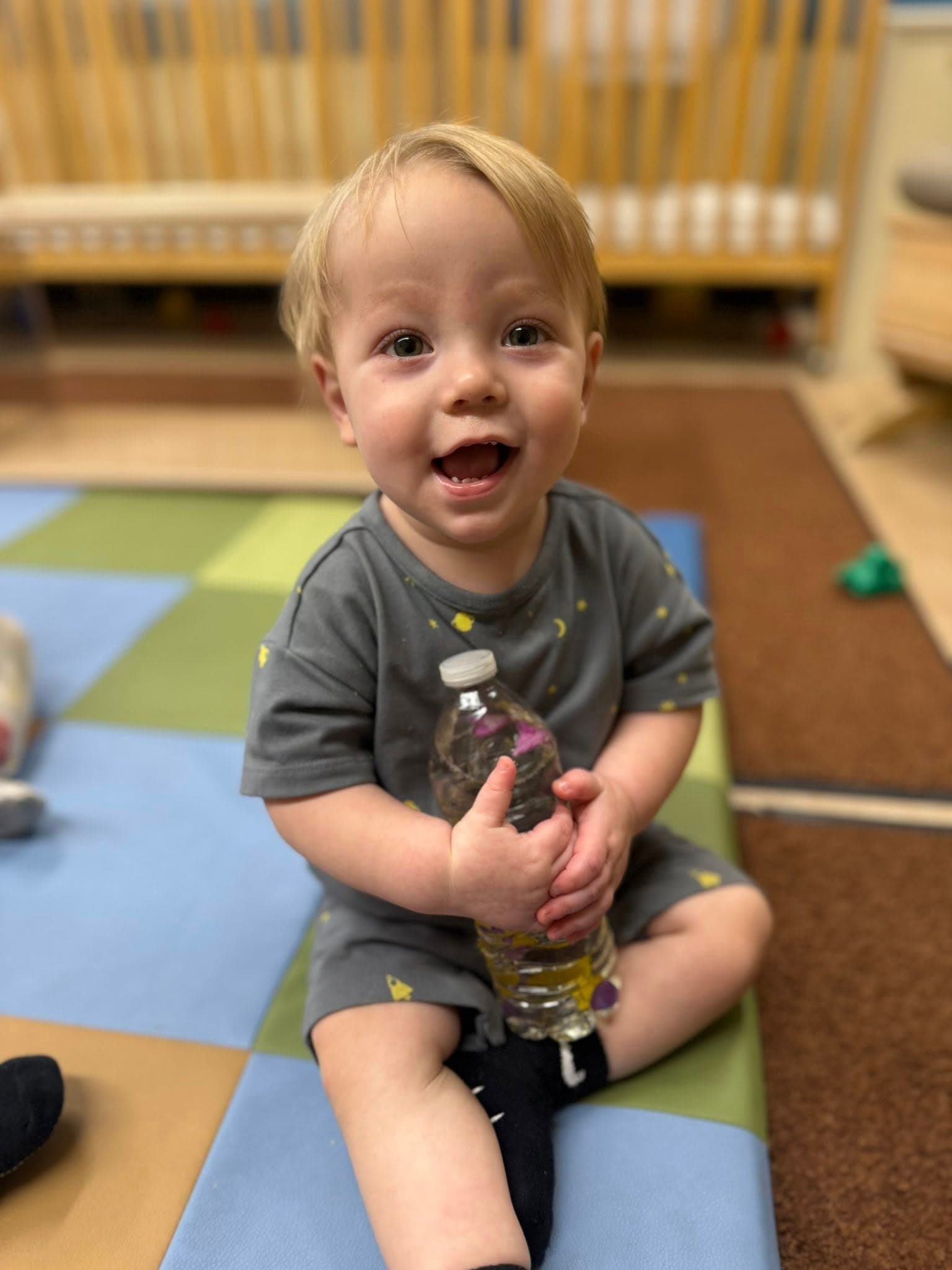 Toddler holding a sensory bottle and smiling during a hands-on learning experience in the infant classroom.