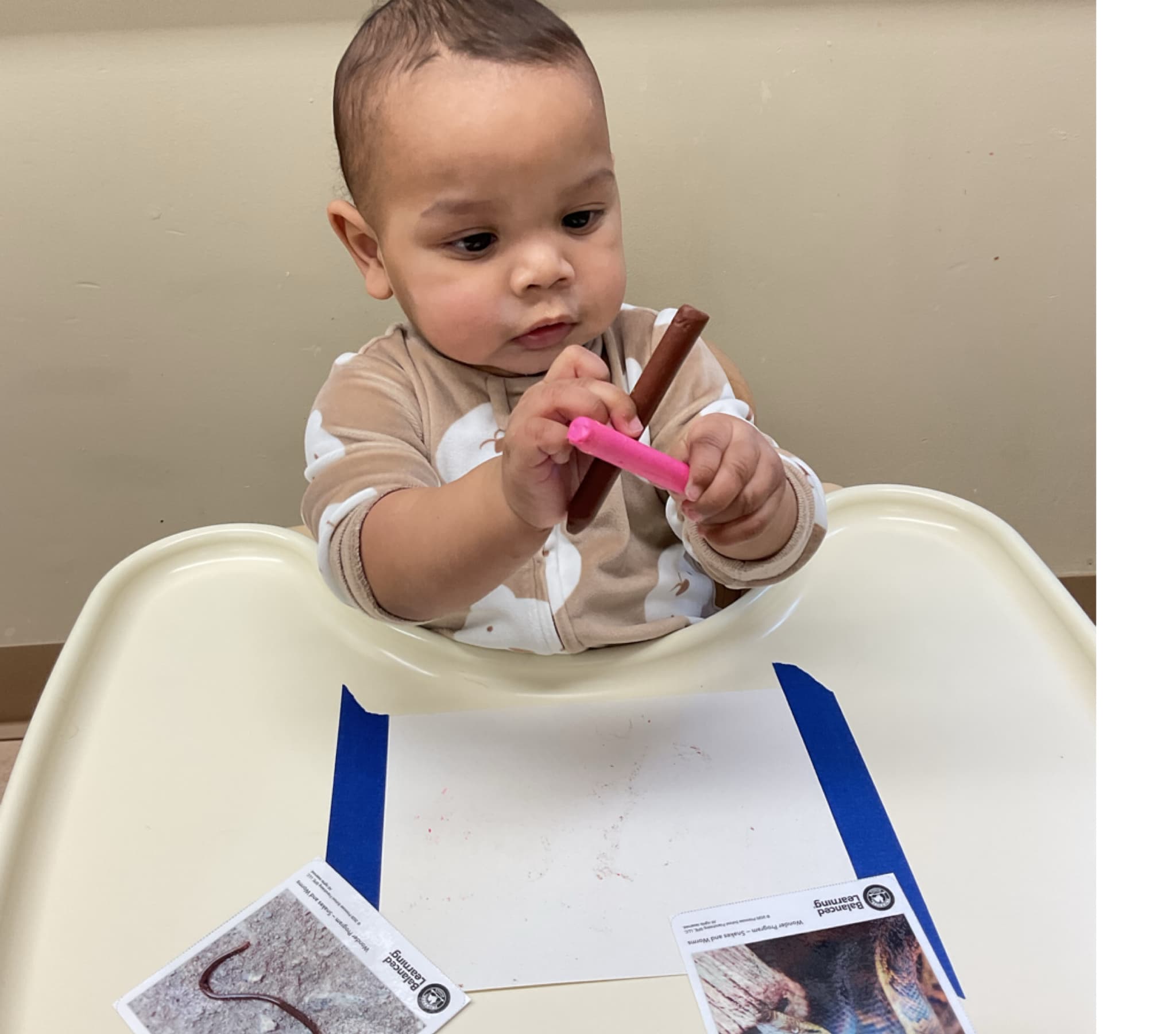 Infant seated in a highchair experimenting with crayons and mark-making during an early art and literacy activity.