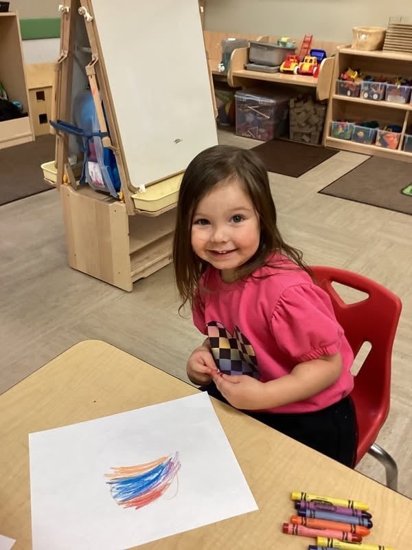 Smiling preschooler creating a colorful drawing with crayons during a classroom activity that builds creativity, fine motor skills, and early writing development at Primrose School.