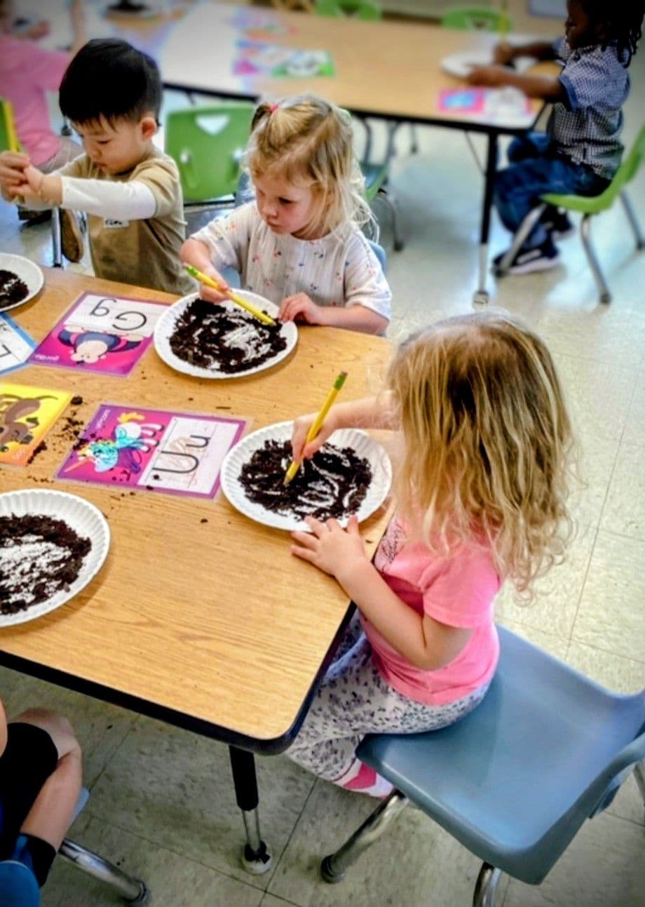 Preschool children practicing letter formation by tracing letters in sensory materials during a hands-on early literacy activity.