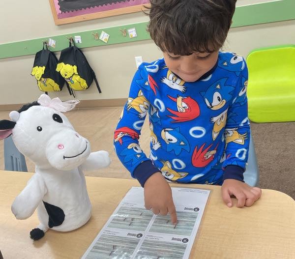 Preschool student pointing to learning materials while participating in a literacy activity alongside a Primrose classroom puppet.