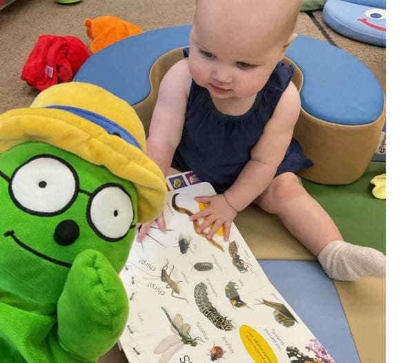 Infant examining an educational picture book alongside a classroom puppet during a guided learning experience.