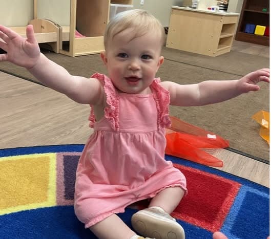 Infant in a pink dress sitting on a colorful classroom rug with arms outstretched, building confidence, balance, and social engagement during playtime.