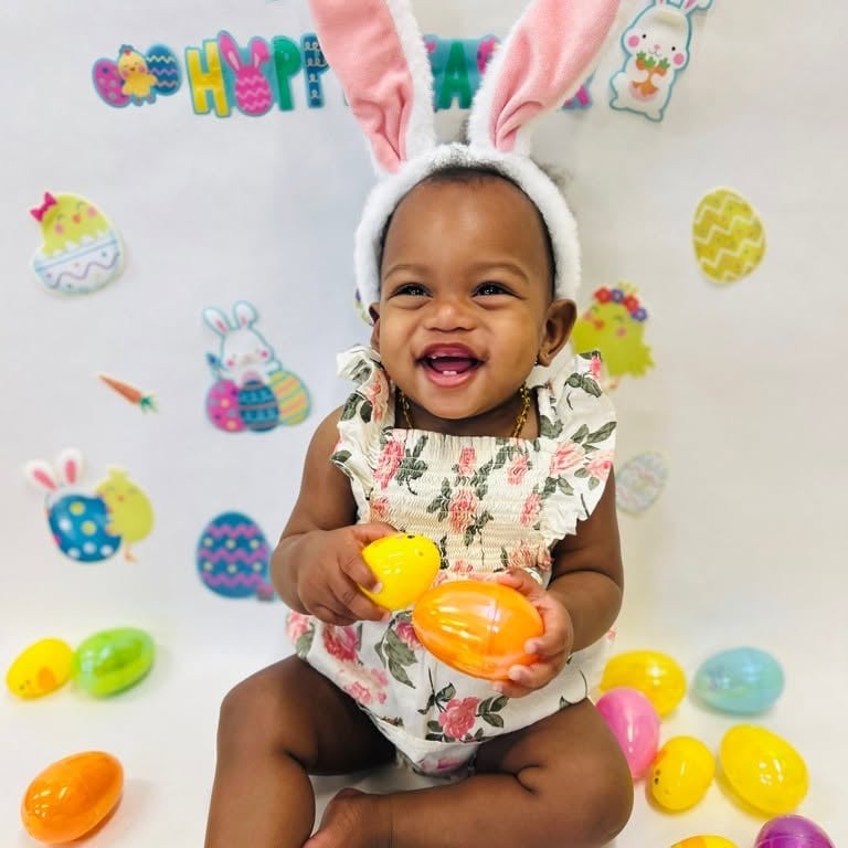 Smiling infant wearing bunny ears while holding colorful Easter eggs during a festive sensory activity that promotes exploration, fine motor skills, and joyful learning at Primrose School.