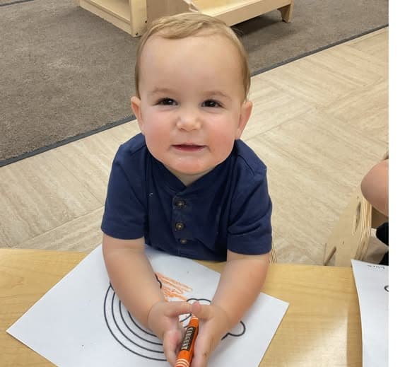 Toddler smiling while coloring on a worksheet and practicing early fine motor and creative skills.