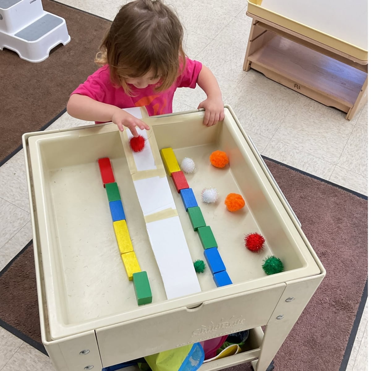Toddler exploring cause-and-effect by guiding colorful pom-poms down ramps while developing fine motor skills, problem-solving, and early STEM concepts through hands-on sensory play at Primrose School.
