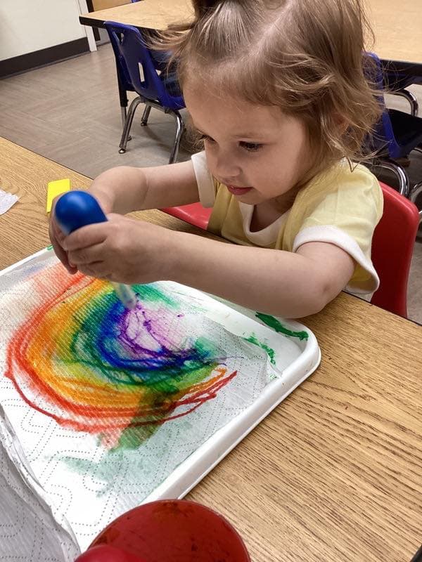 Toddler using paint tools to blend vibrant colors into a rainbow-inspired art project during a sensory learning activity.