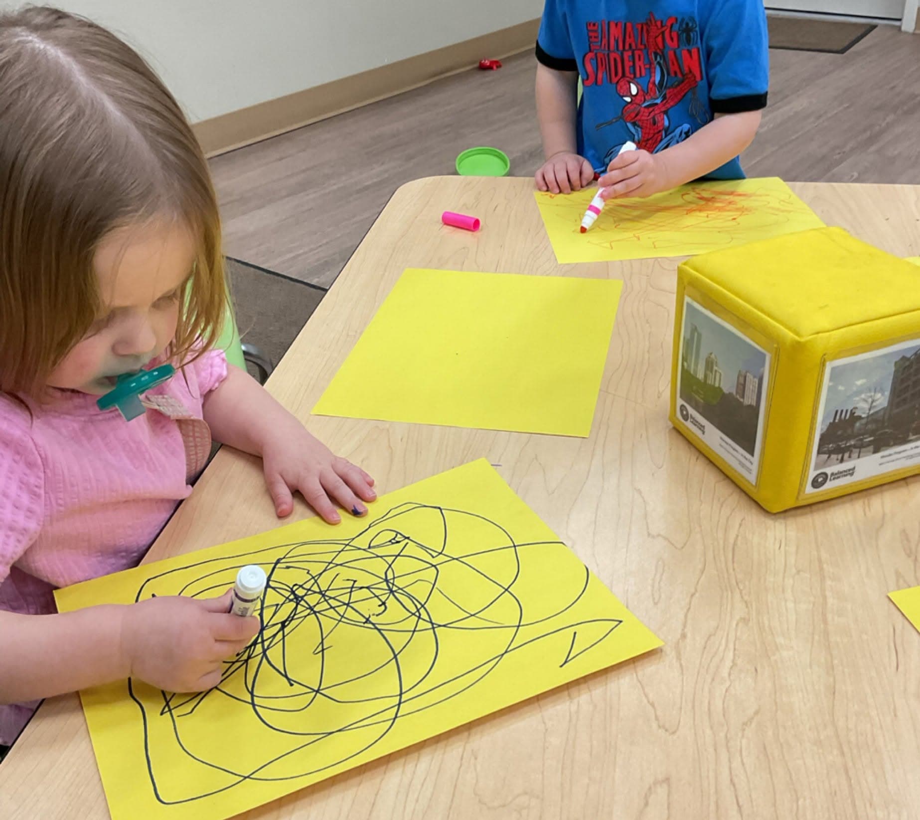 Young child drawing with markers on yellow paper during a classroom art activity that encourages creativity, fine motor development, and self-expression.