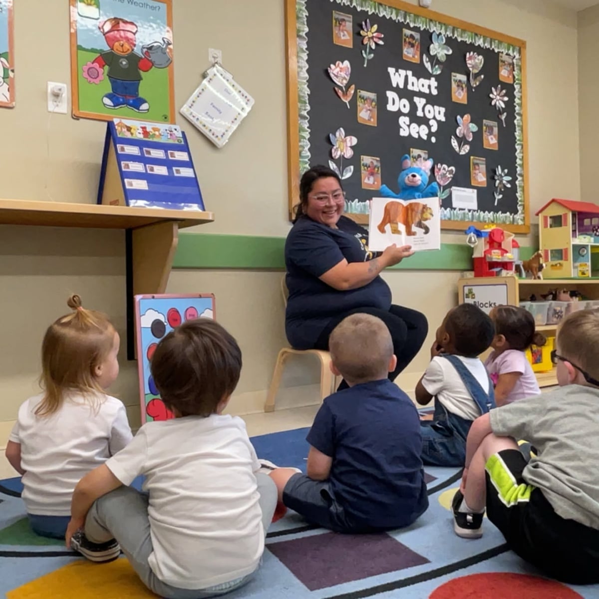Teacher reading a story aloud to preschool students during interactive group time, encouraging listening skills, vocabulary development, comprehension, and a love of books at Primrose School.