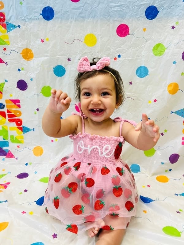 Smiling infant wearing a strawberry-themed birthday outfit while celebrating a first birthday milestone at Primrose School with a festive backdrop.