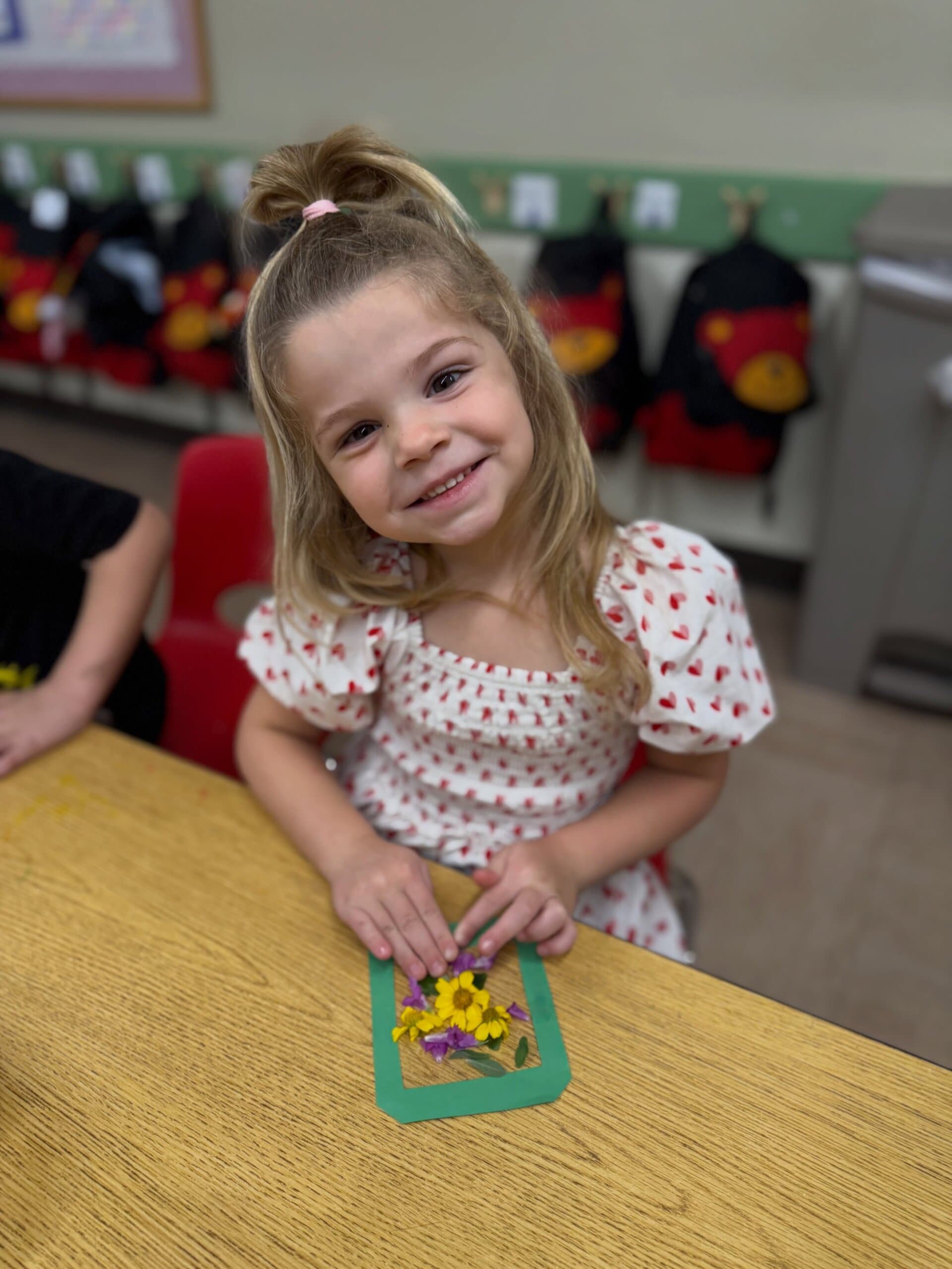 Smiling preschool student proudly displaying a handmade flower art project created with colorful pressed flowers and natural materials.