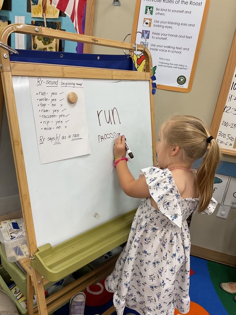 Preschool student practicing beginning letter sounds and writing words on a classroom whiteboard to strengthen early reading and phonics skills.