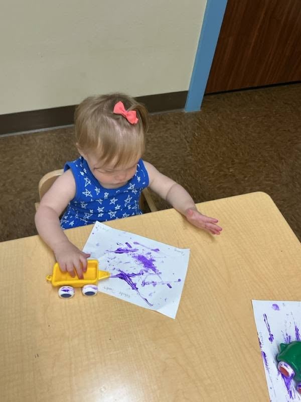 Young toddler creating colorful artwork by rolling a toy truck through paint, encouraging sensory exploration, fine motor development, and creativity at Primrose School.