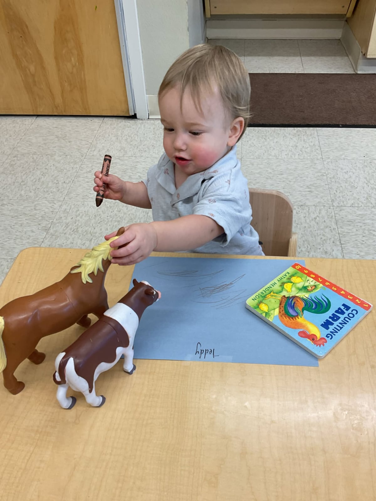 Toddler exploring toy farm animals alongside a farm-themed book and drawing activity to build vocabulary, creativity, fine motor skills, and imaginative play at Primrose School.