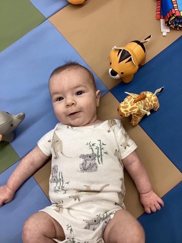 Smiling infant lying on a colorful classroom mat surrounded by soft animal toys during sensory exploration at Primrose School