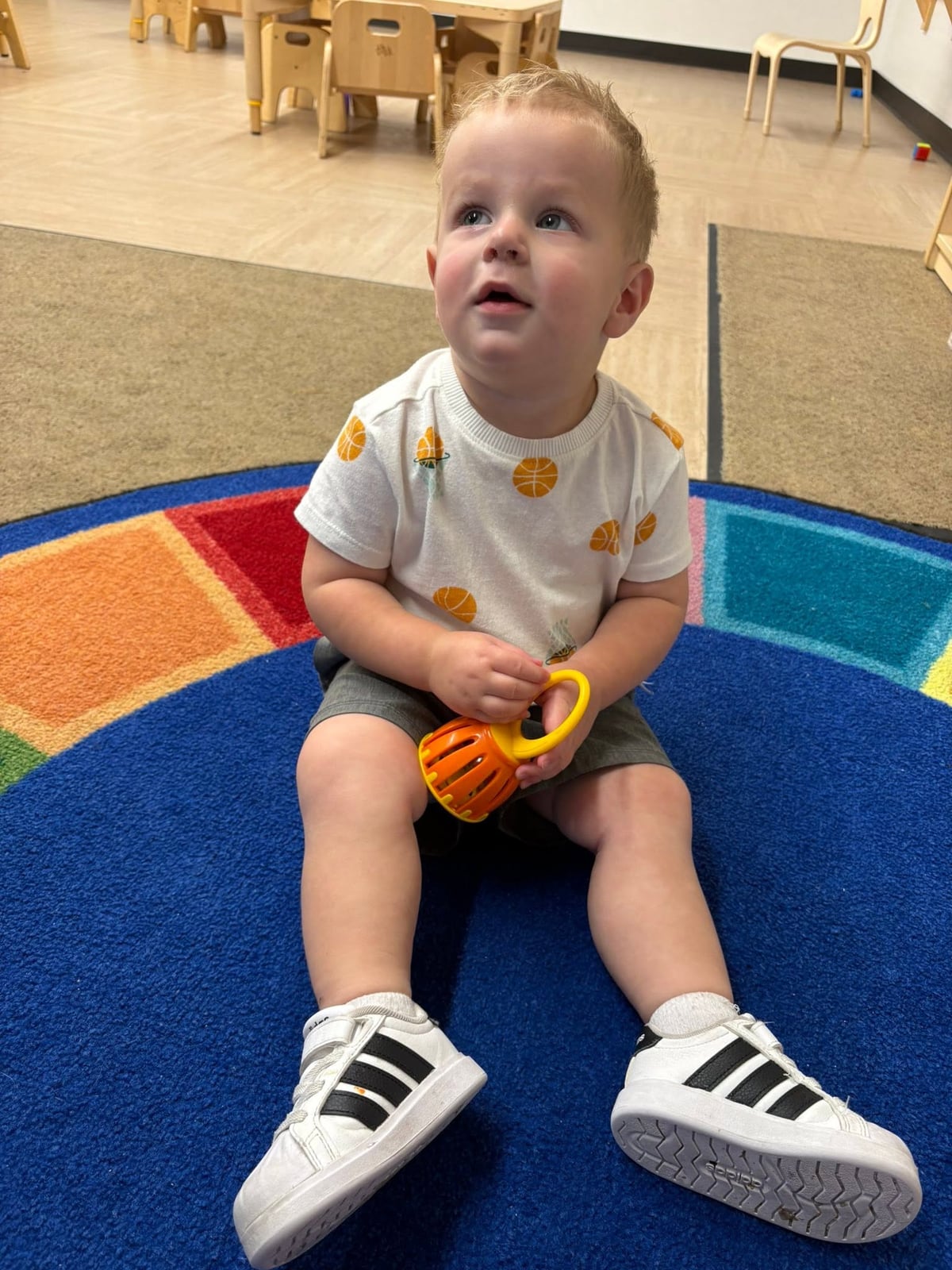 Toddler sitting on a colorful classroom rug while holding a musical rattle during an activity that encourages sensory exploration and motor skill development at Primrose School.
