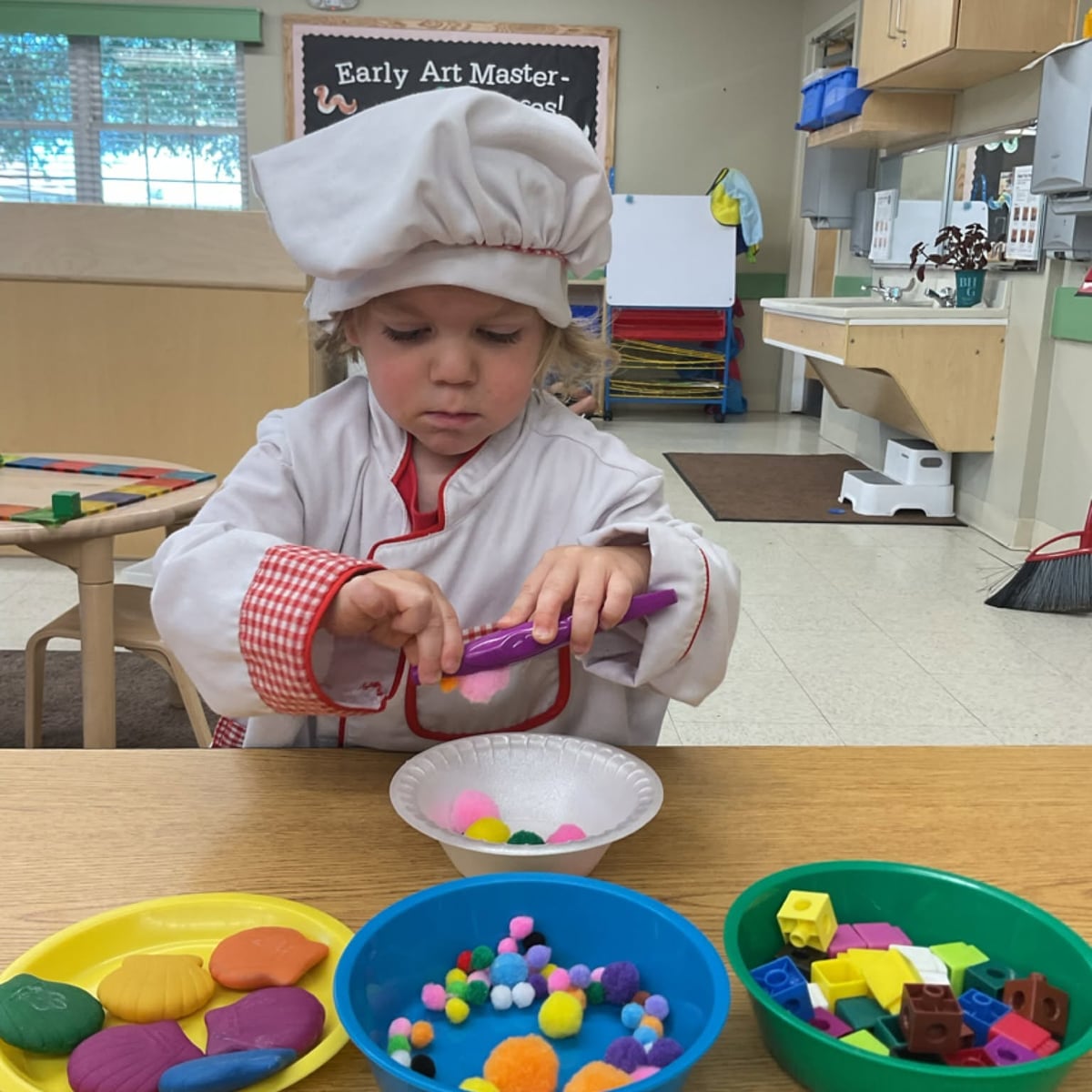 Preschool student dressed as a chef using classroom manipulatives to strengthen fine motor skills, creativity, counting, and imaginative role-play during hands-on learning at Primrose School.