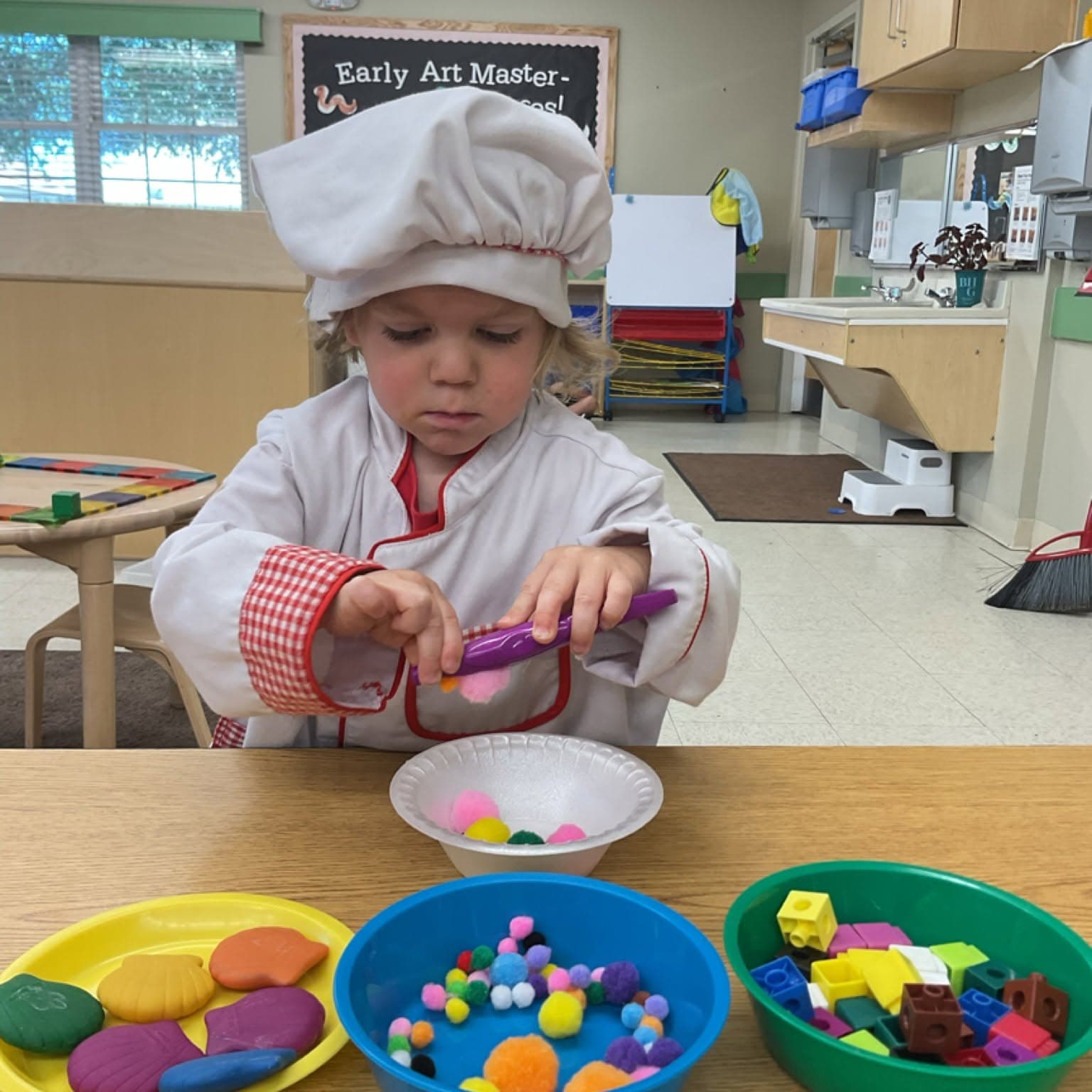 Preschool student dressed as a chef using classroom manipulatives to strengthen fine motor skills, creativity, counting, and imaginative role-play during hands-on learning at Primrose School.
