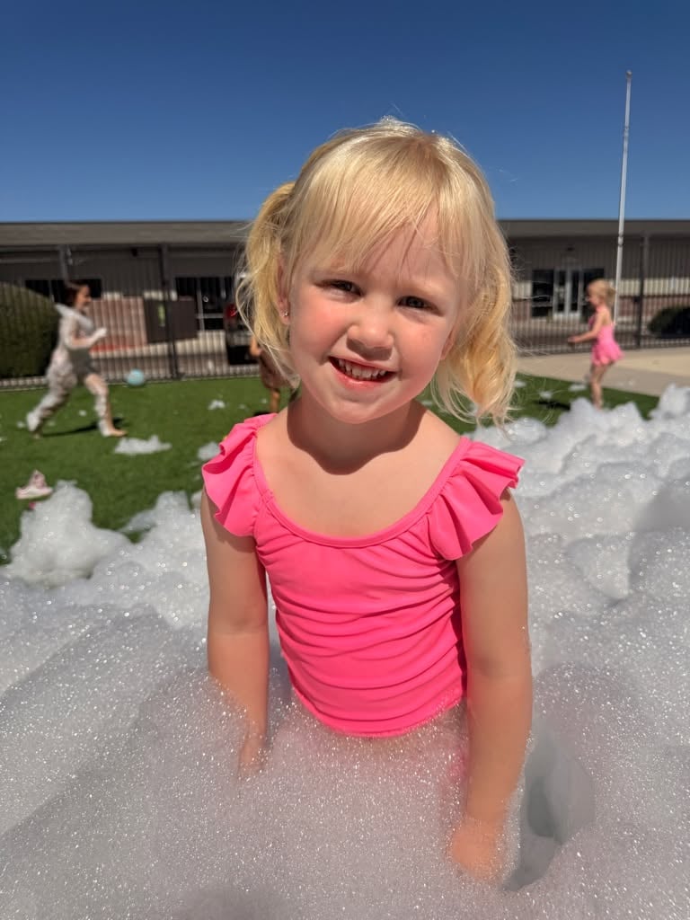 A smiling preschool student stands in a mountain of foam during an outdoor Foam Party at Primrose School while classmates play in the background, enjoying a fun-filled summer activity.