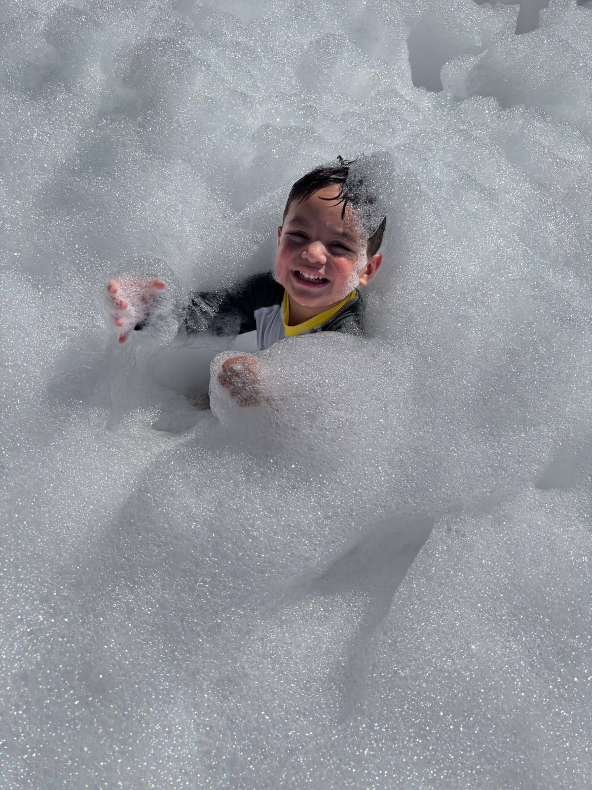 A smiling preschool student stands in a mountain of foam during an outdoor Foam Party at Primrose School while classmates play in the background, enjoying a fun-filled summer activity.