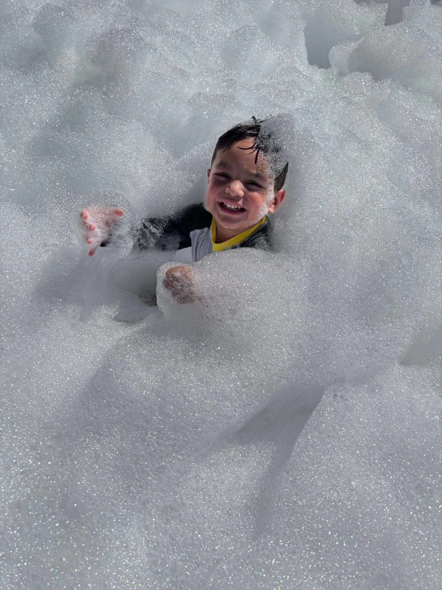 A smiling preschool student stands in a mountain of foam during an outdoor Foam Party at Primrose School while classmates play in the background, enjoying a fun-filled summer activity.