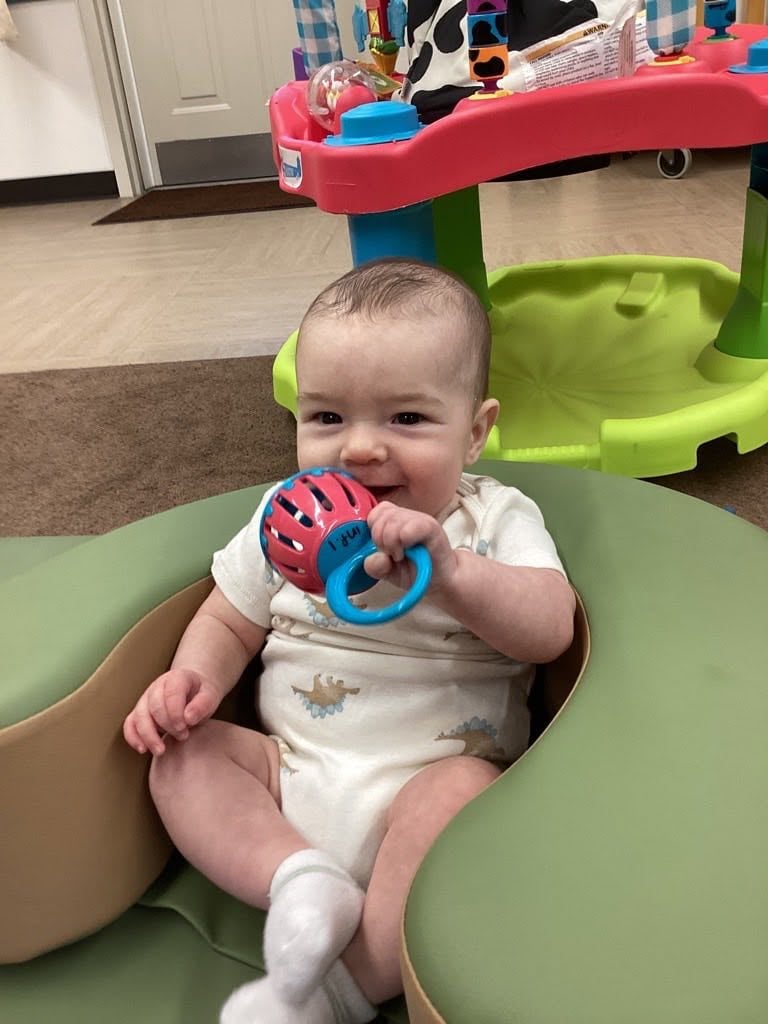 Smiling infant sitting in a supportive play seat while holding a colorful rattle during interactive play at Primrose School.