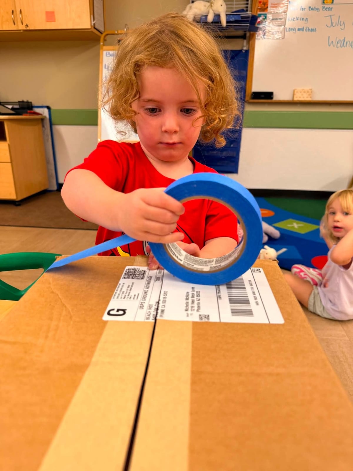 Preschooler helping tape a cardboard box during a hands-on classroom activity that encourages teamwork, problem-solving, and fine motor development at Primrose School.