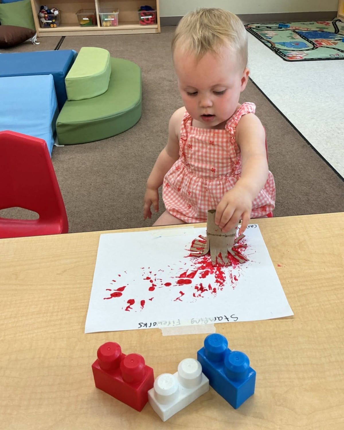 Toddler using a cardboard tube to stamp colorful paint and create firework-inspired artwork during a hands-on classroom art activity at Primrose School.