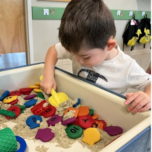 Preschool student exploring a sensory bin filled with numbered manipulatives while strengthening number recognition, fine motor skills, sorting, and early math concepts through hands-on learning at Primrose School.
