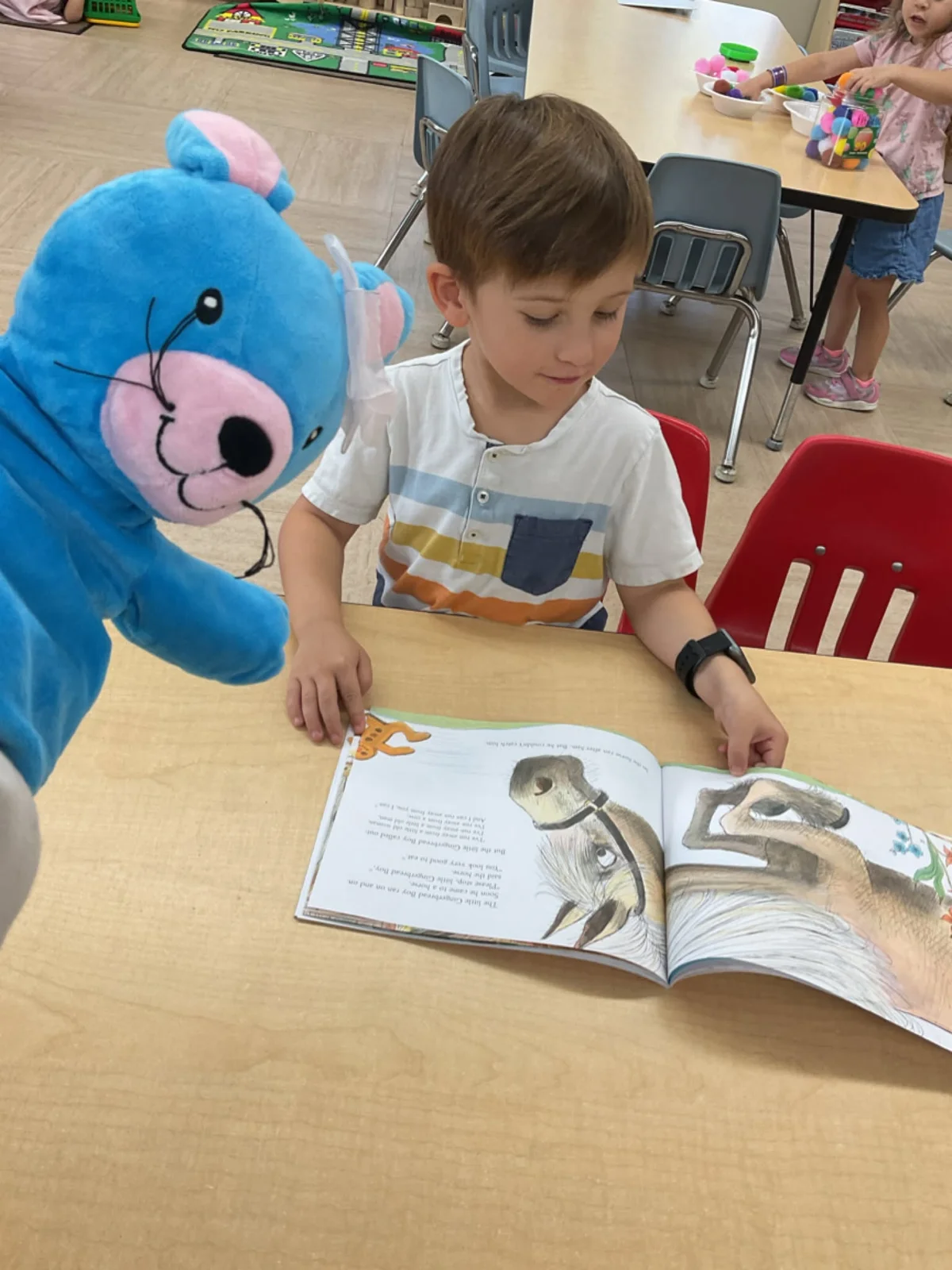 A preschool student reads a picture book alongside Primrose Friend Benjamin the Bear during a literacy activity, encouraging early reading skills and a love of books.