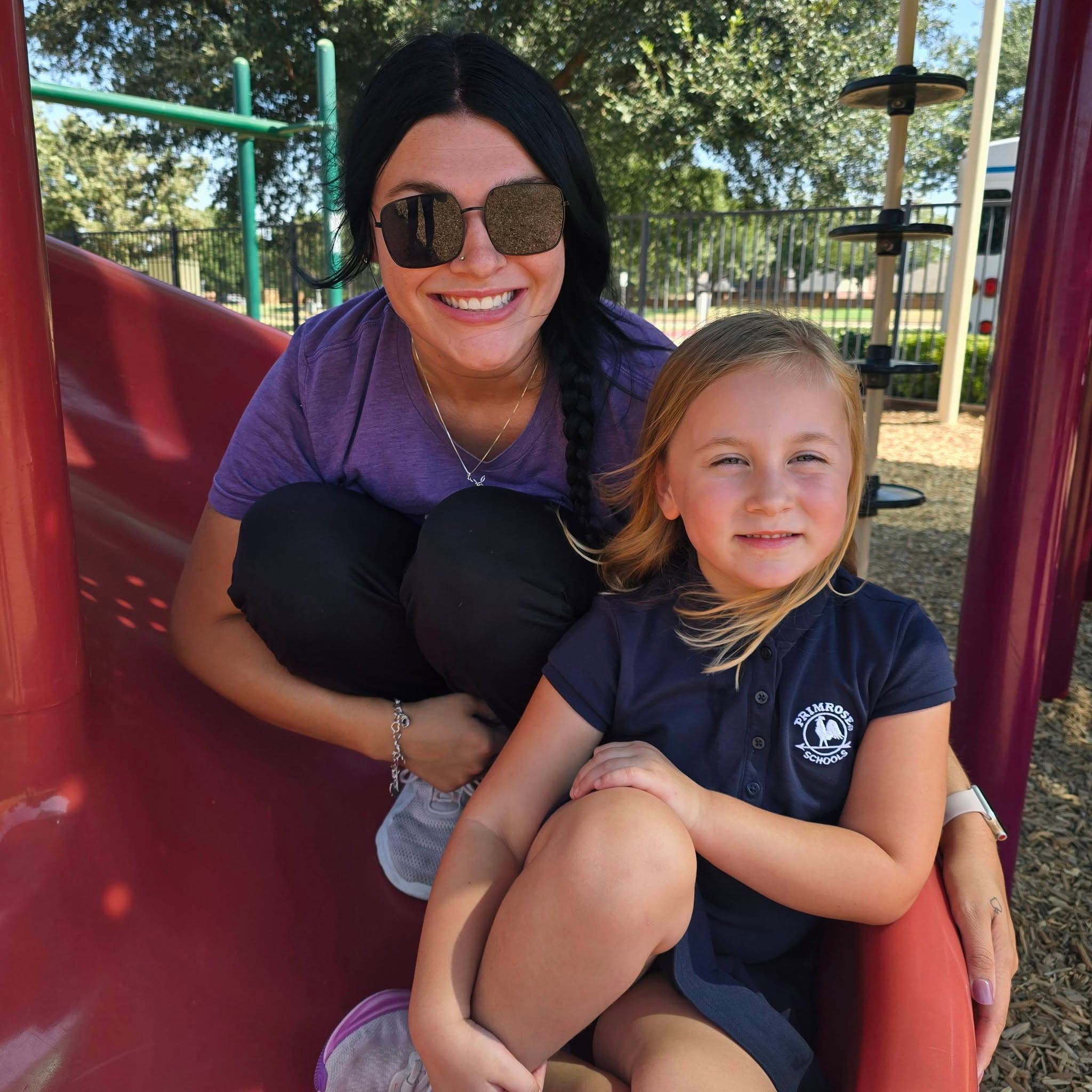 A Pre-Kindergarten teacher sitting on the slide with one of her students at Primrose School at Heritage in Fort Worth, TX.