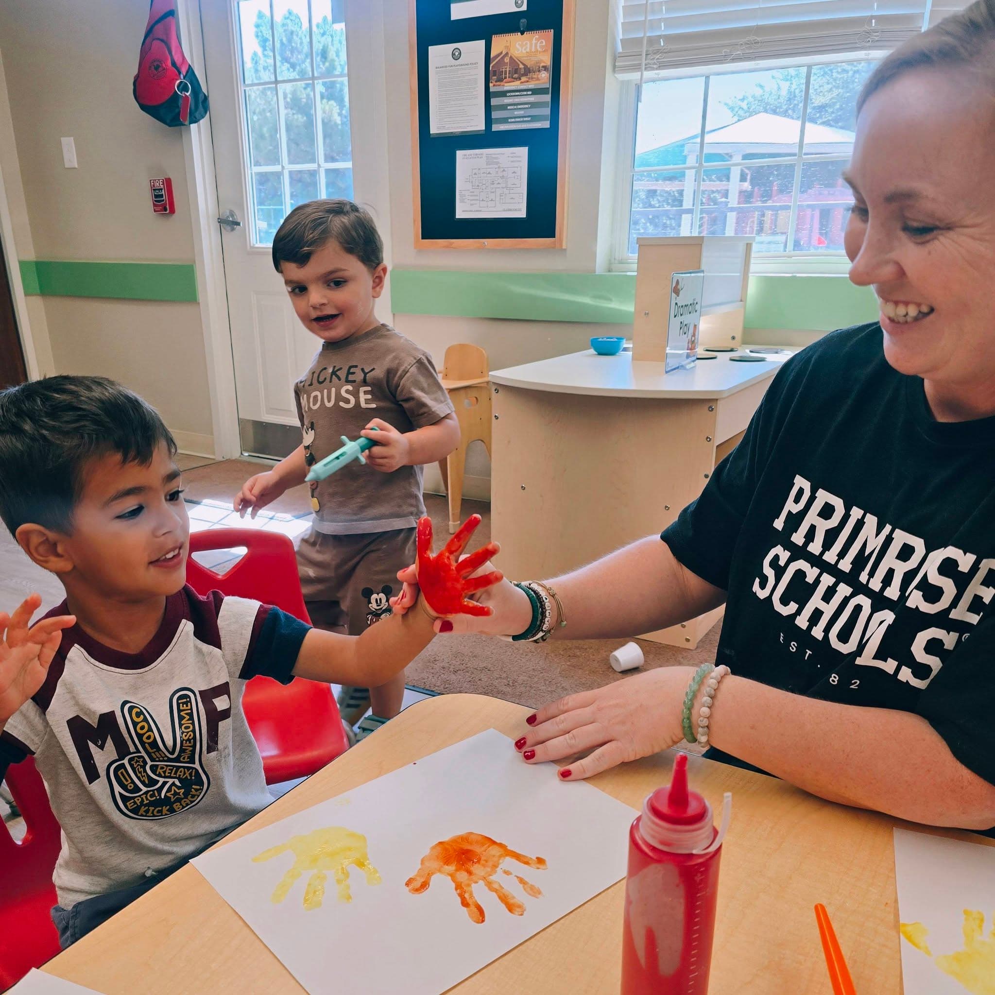 A preschool pathways teacher helping a smiling child in her class to place a handprint on paper at Primrose School at Heritage in Fort Worth, TX.