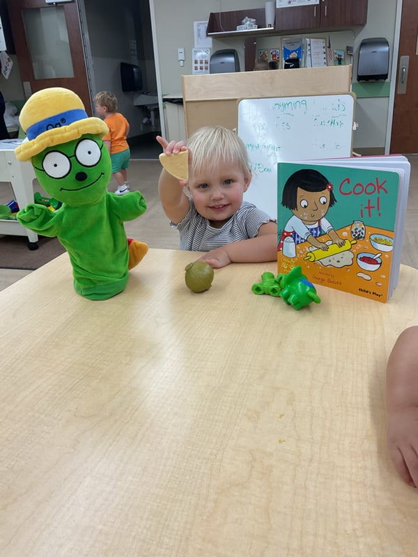 Toddler exploring pretend food and reading the book Cook It! while engaging in imaginative play with a classroom character puppet.