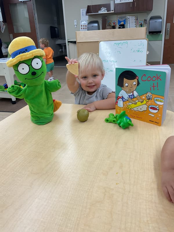 Toddler exploring pretend food and reading the book Cook It! while engaging in imaginative play with a classroom character puppet.