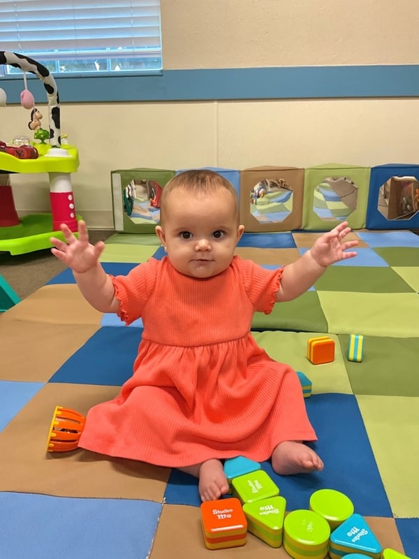 A baby in an orange dress with her arms raised up surrounded by musical shaker toys.