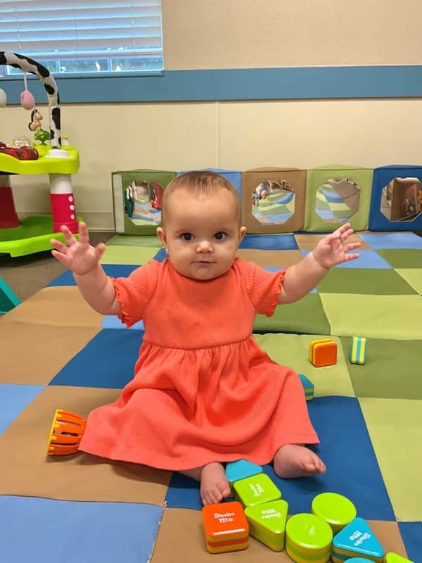 A baby in an orange dress with her arms raised up surrounded by musical shaker toys.