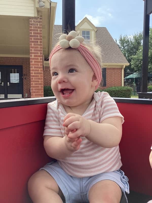 A baby girl with a pink flower bow riding in a red train car, with a happy smile on her face.