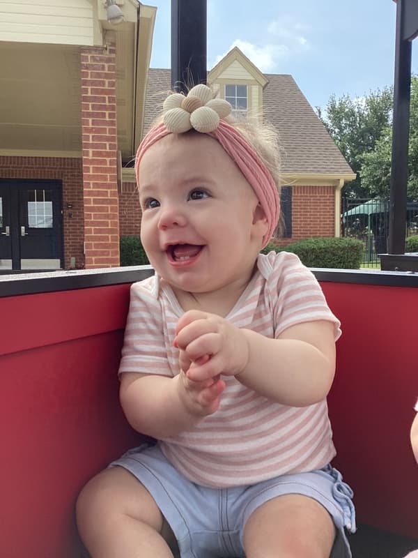A baby girl with a pink flower bow riding in a red train car, with a happy smile on her face.
