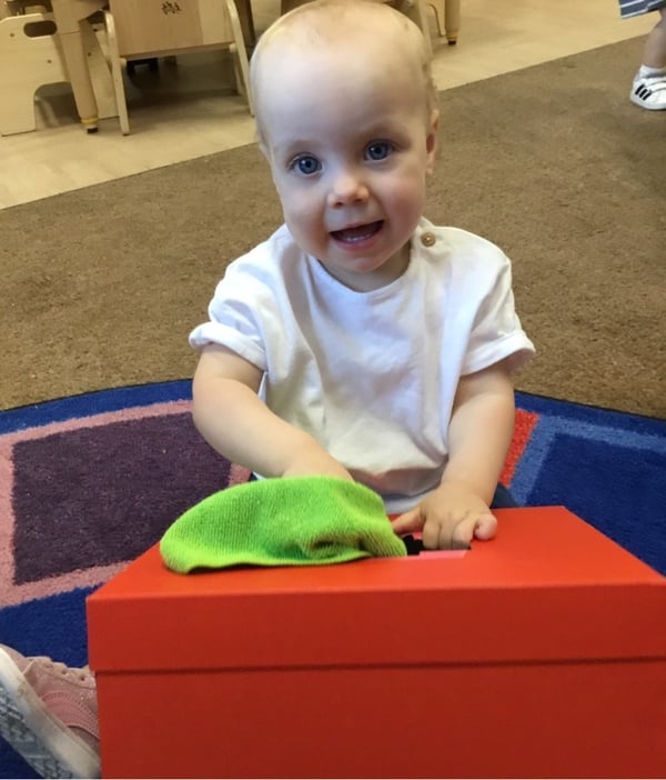 Smiling toddler exploring putting a scarf in a box in our toddler classroom