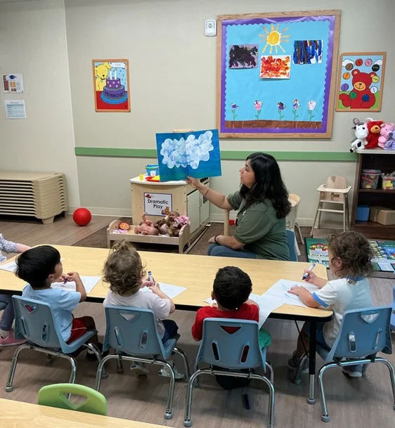 A teacher reading a book about clouds while children sit at the table with crayons and paper